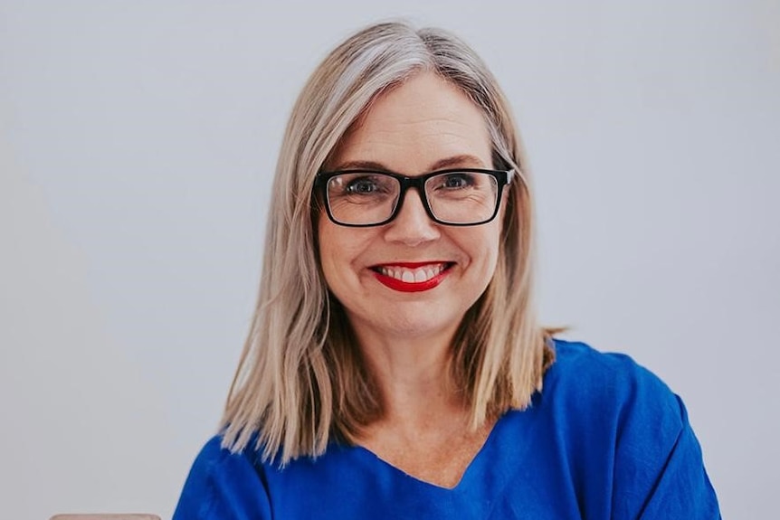 A woman in her early 50s with blonde/gray hair, dark glasses, red lipstick, blue shirt, smiling