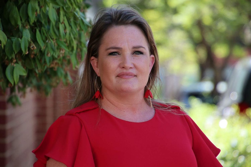 Tanya Steinbeck wearing a red shirt standing in front of green bushes and a brick wall.