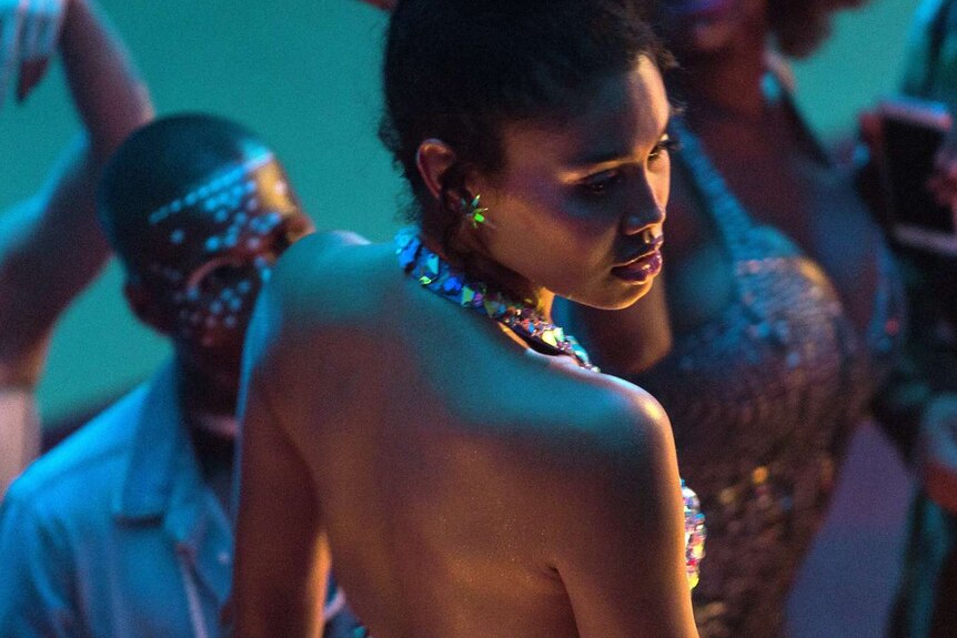 Blue-lit shot from above and behind, looking down on profile of young woman with bare back and shoulders, surrounded by people.