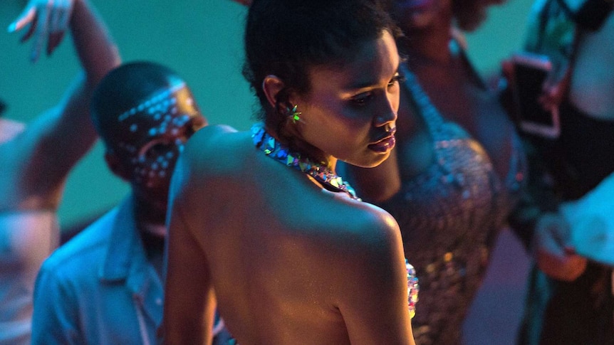 Blue-lit shot from above and behind, looking down on profile of young woman with bare back and shoulders, surrounded by people.