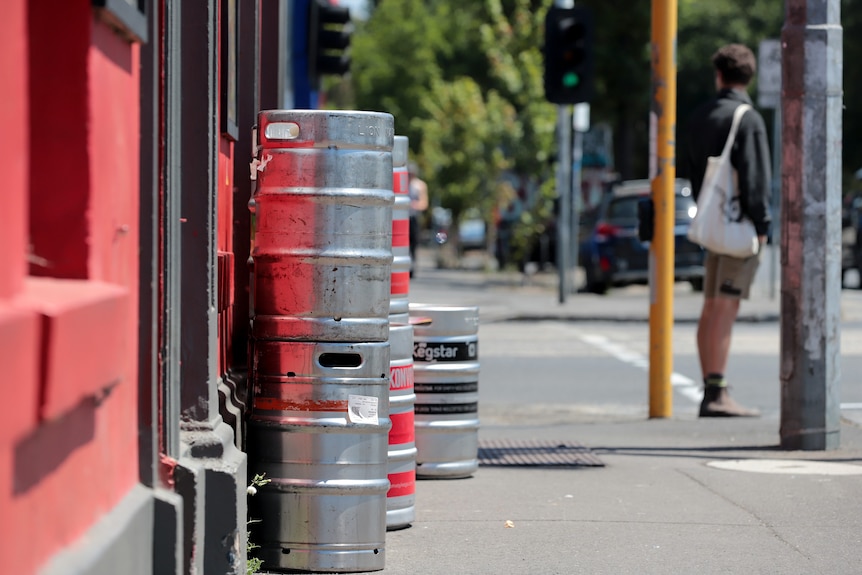 Una fila de barriles de cerveza plateados afuera de un pub de paredes rojas con una intersección al fondo donde espera un hombre