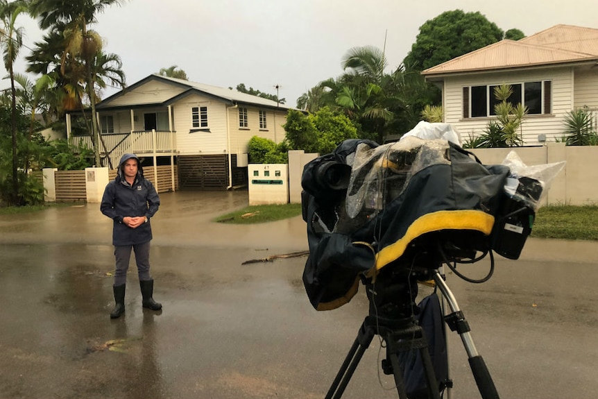 Bavas standing in raincoat and gumboots in front of camera filming flooded homes