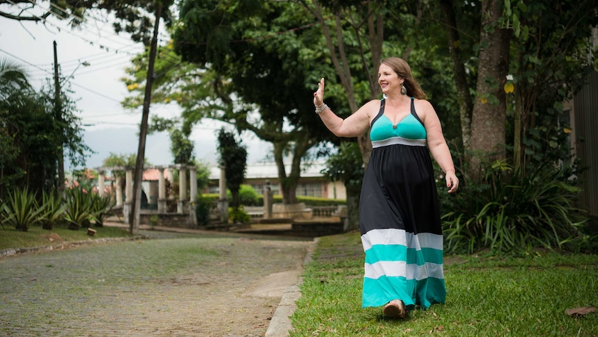 waving woman in a long black and aqua dress walks along a street with trees in the background