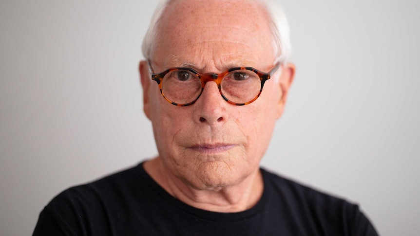 A man with white hair and glasses looks at the camera.