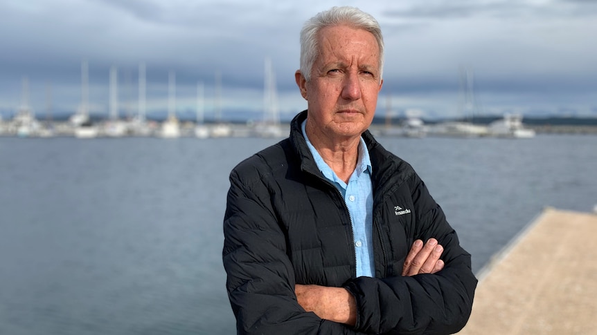 Man wearing jacket and shirt standing at a boat harbour with arms crossed