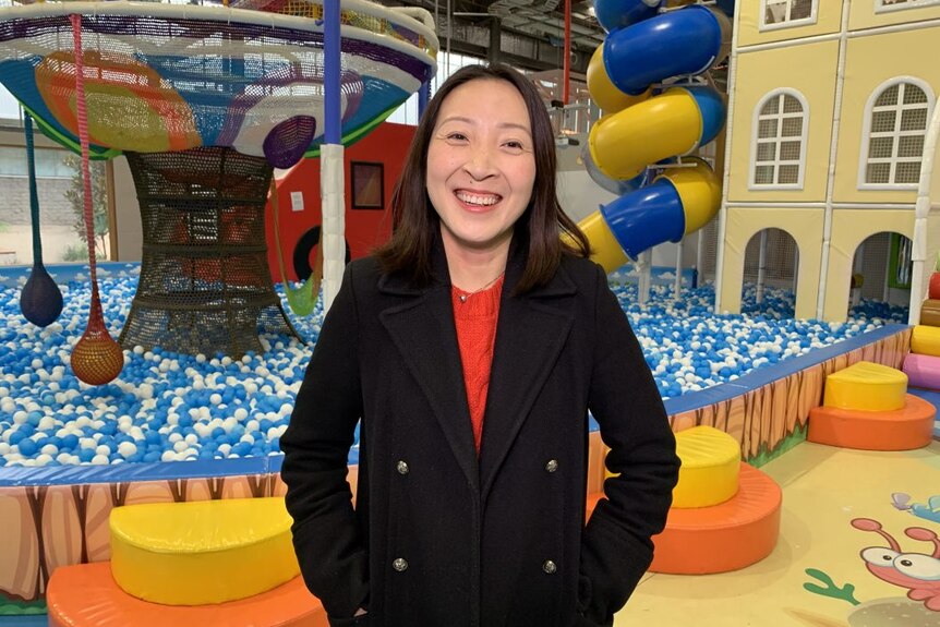 A woman smiles at the camera. Behind her is an enormous ball pit with slides and play equipment rising out of it.