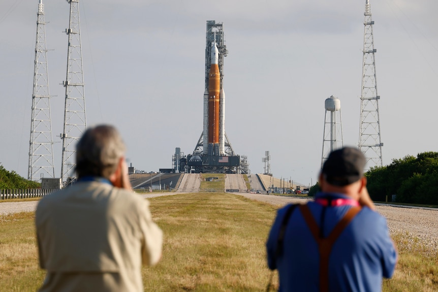 two people stand in front of a rocket at a launch pad
