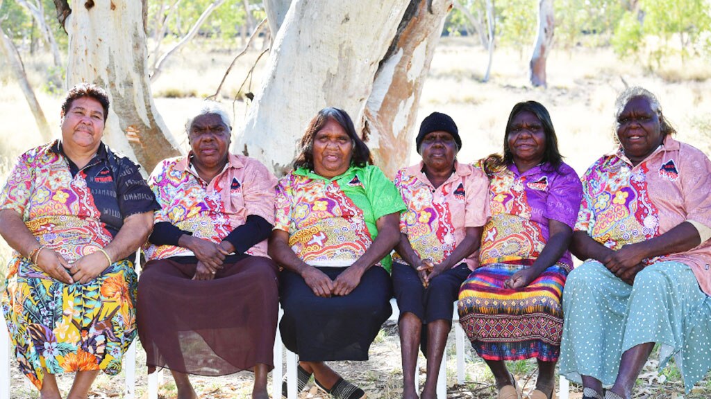 A group of middle-aged Aboriginal women sit in plastic chairs looking at the camera.