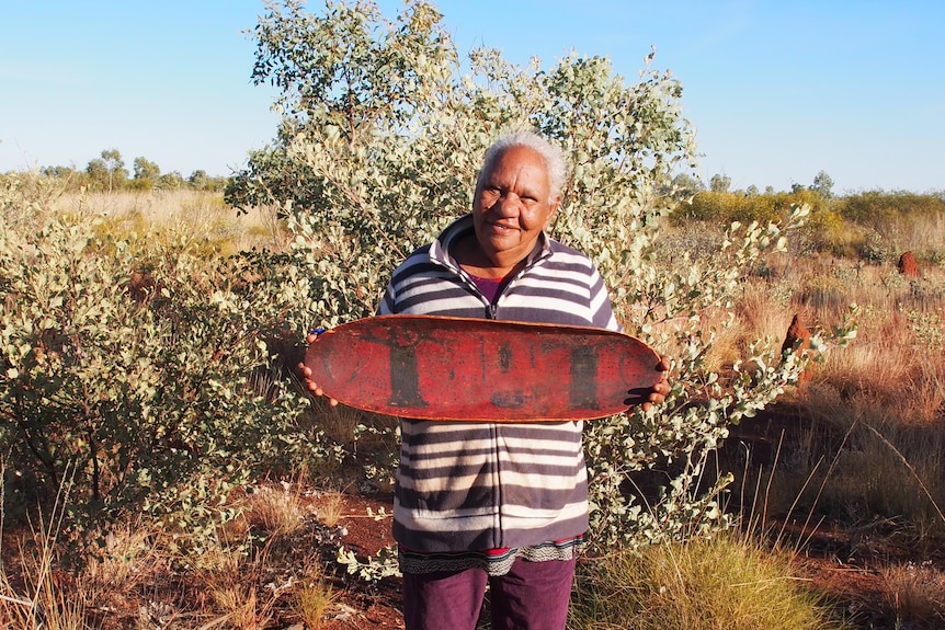 An indigenous person holding a red-coloured Coolamon, a long shallow item, decorated with black images of boab trees