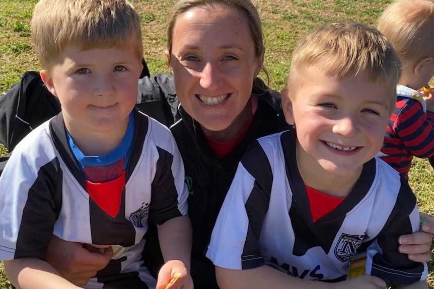 A mum smiles with her arms around two young boys in sports uniforms.