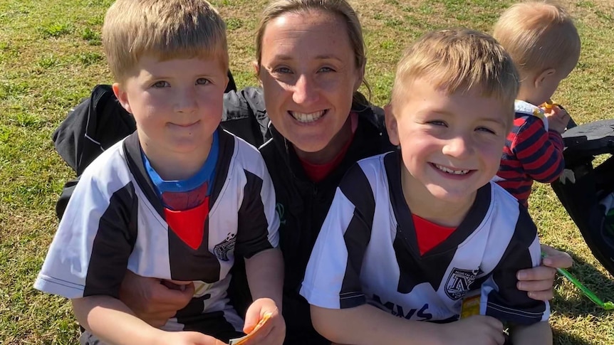 A mum smiles with her arms around two young boys in sports uniforms.