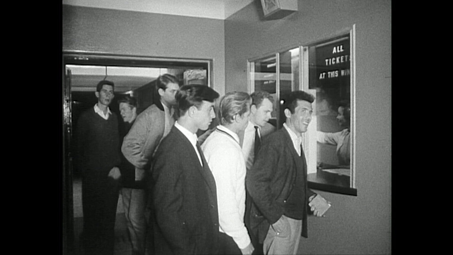 Una foto en blanco y negro de los cineastas sonrientes sonrientes haciendo cola en una taquilla en un cine de Sydney en la década de 1960. 