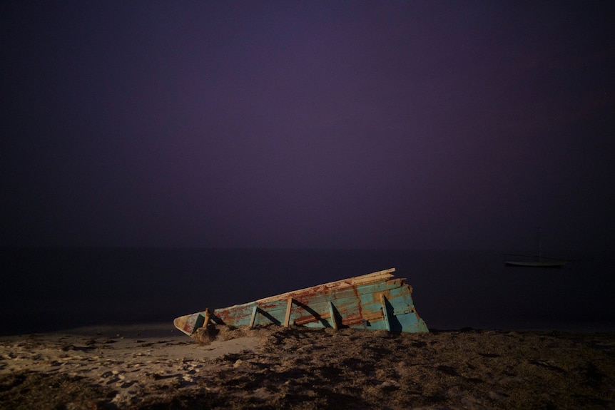The wreck of a traditional Mauritanian fishing boat lies on a beach at dusk.