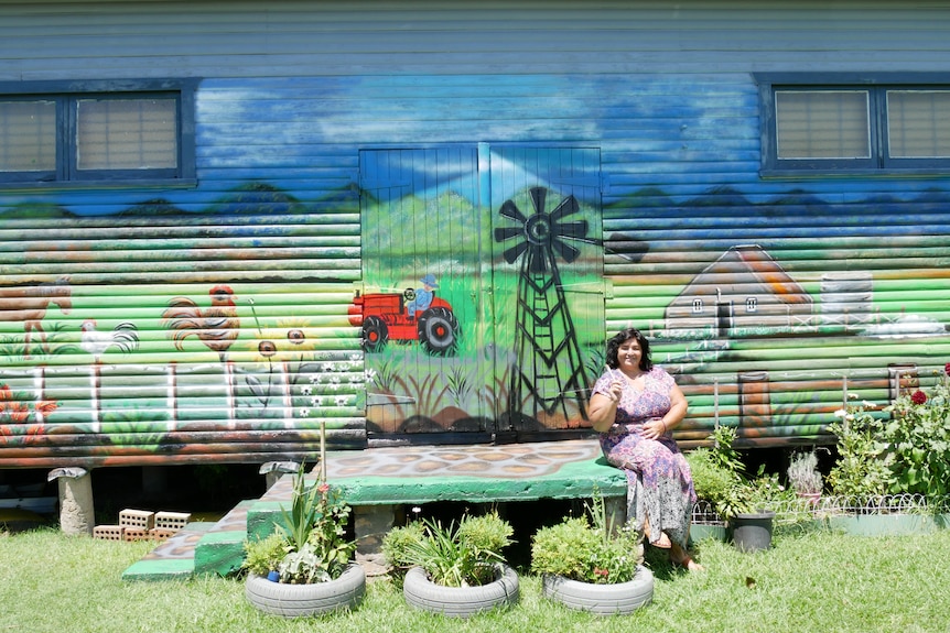 Woman in a colourful dress sits on a bench in front of a bright mural along a building wall.