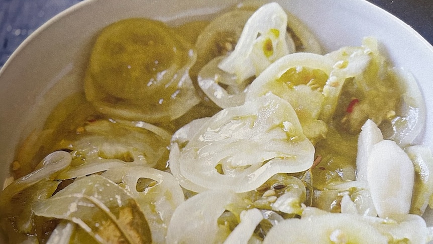 Sliced pickled green tomatoes and bay leaves in a white bowl on a blue table.