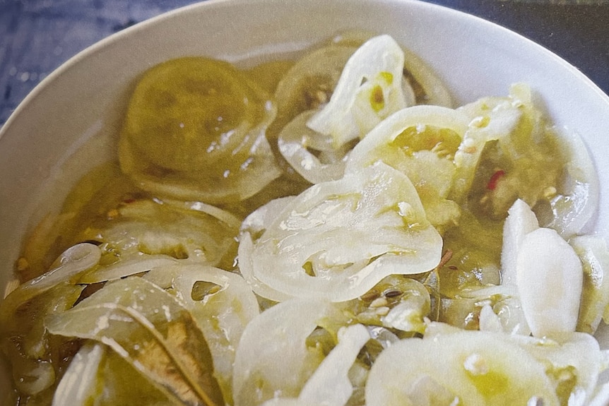 Sliced pickled green tomatoes and bay leaves in a white bowl on a blue table.