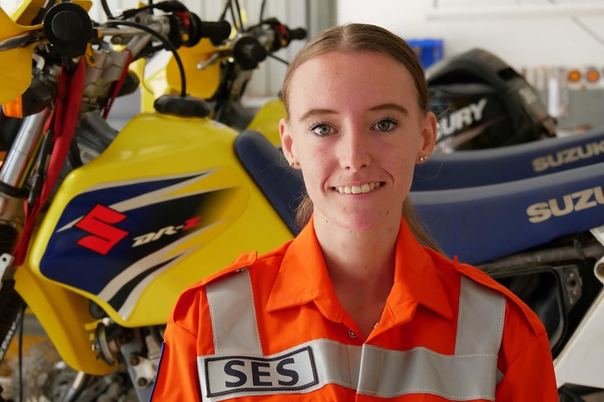 A young women in an SES orange jumpsuit stands in font of a yellow and blue motorbike.