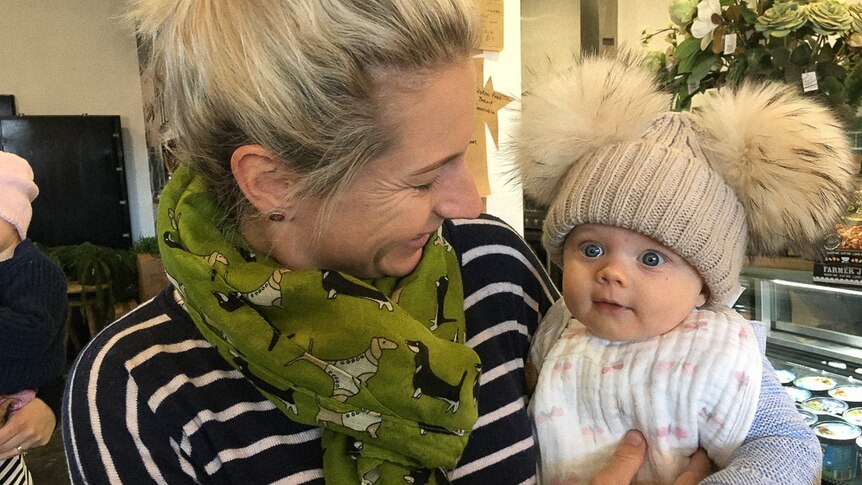 Woman holding baby in cafe, baby wearing beanie with fluffy pom poms