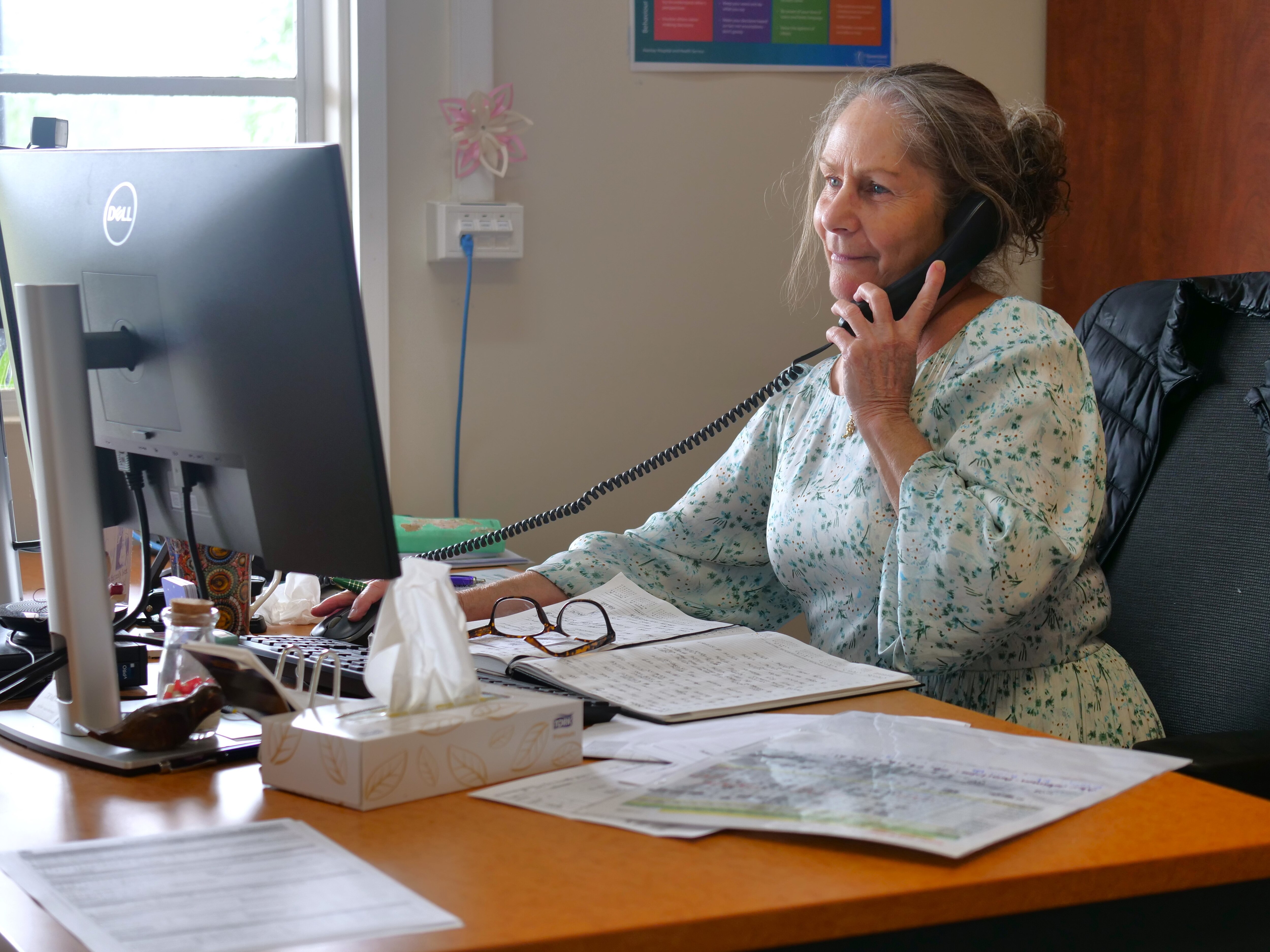 A nurse sits at her desk looking at a computer screen while talking into a landline phone
