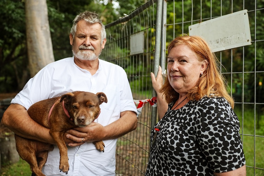 A man and woman, Suzie and Joe Haddock stand at a temporary fence. Suzie is touching the fence and Joe holds their dog Karl Barx