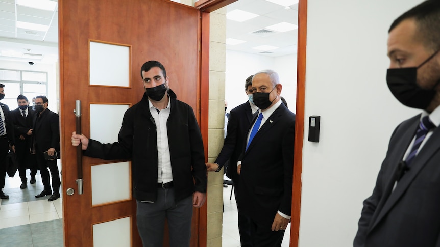 An elderly man in a dark suit walks into a building as a taller man opens the door.