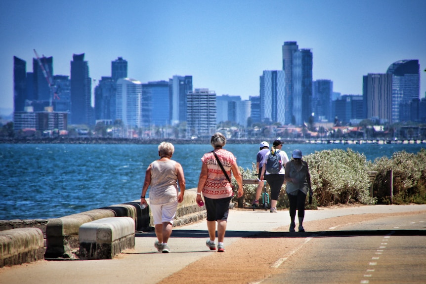People walk along a foreshore in Melbourne, with the city skyline in the back on a sunny day.