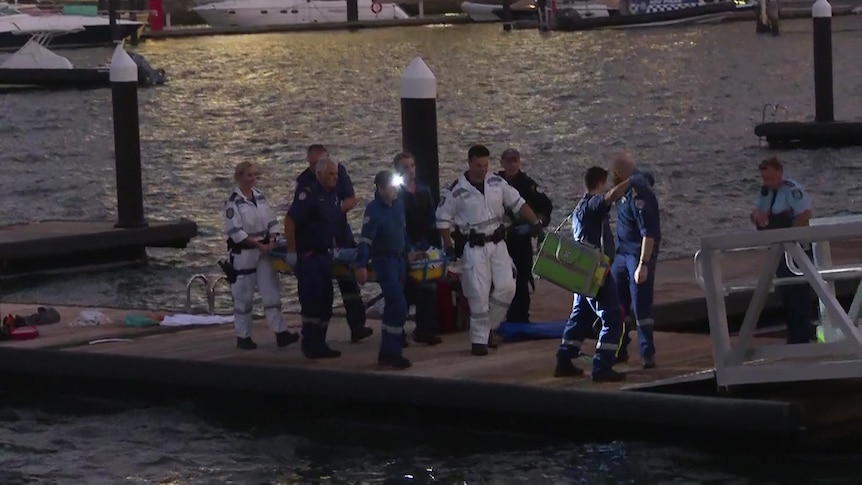 Several emergency workers carry on a woman on a stretcher along a dock.