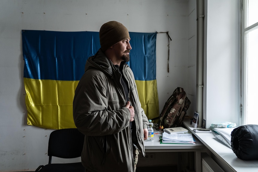 A man in khaki jacket and beanie looks out the window while a Ukrainian flag hangs on the wall behind him