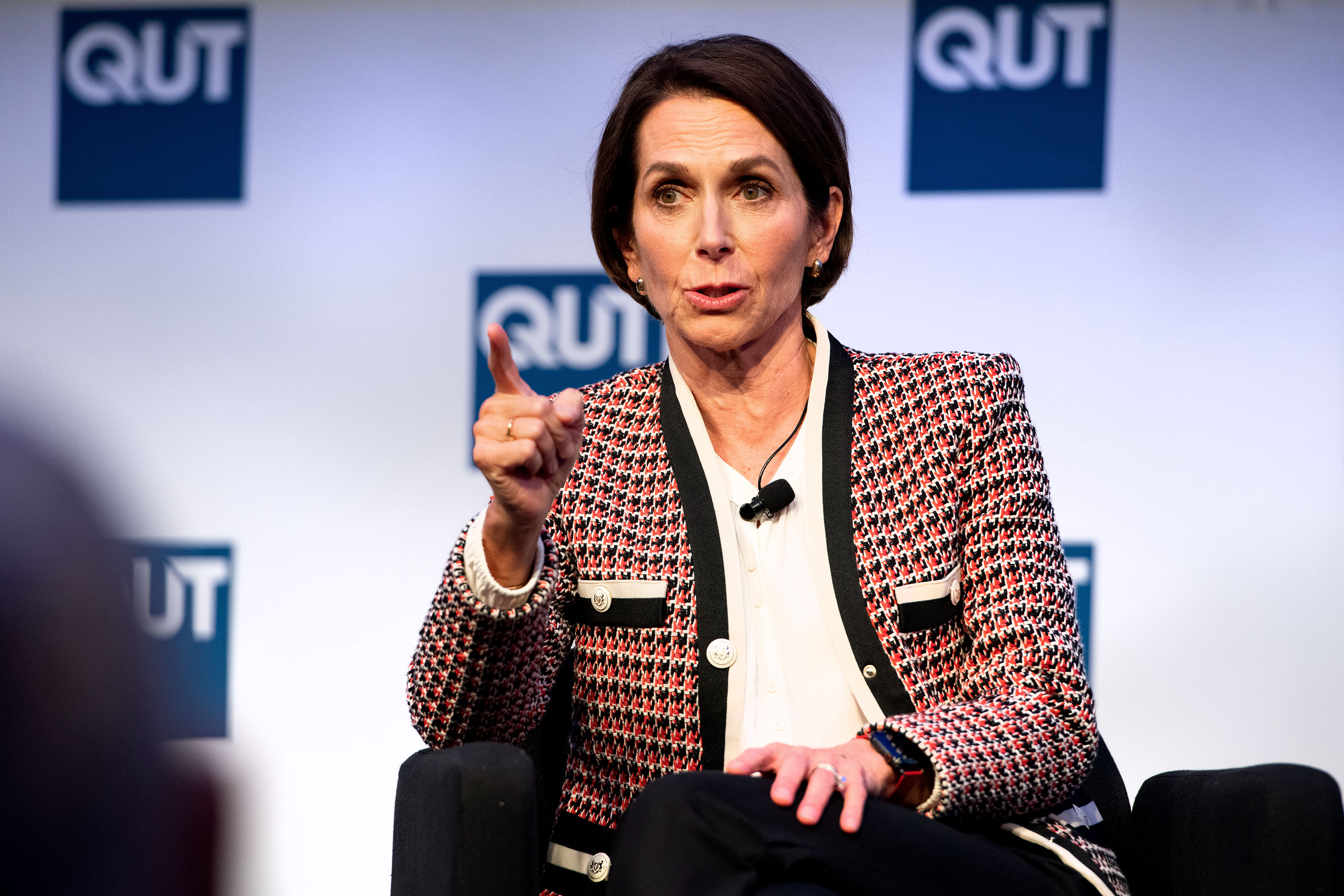 A woman with short brown hair wearing a black and red patterned blazer mid-sentence on a stage pointing her finger.
