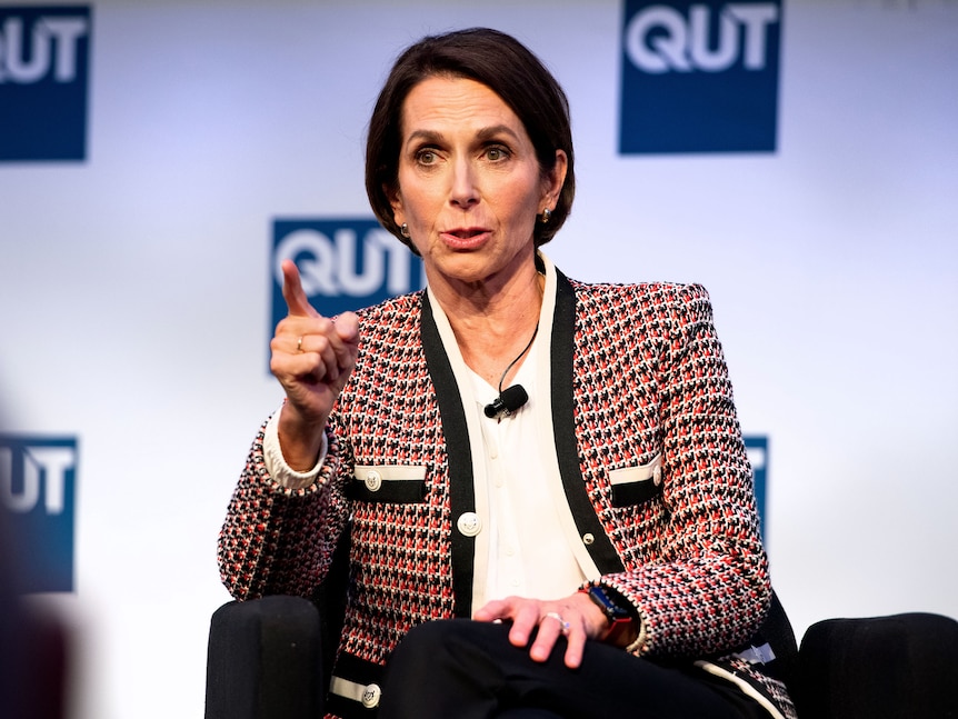 A woman with short brown hair wearing a black and red patterned blazer mid-sentence on a stage pointing her finger.