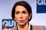 A woman with short brown hair wearing a black and red patterned blazer mid-sentence on a stage pointing her finger.