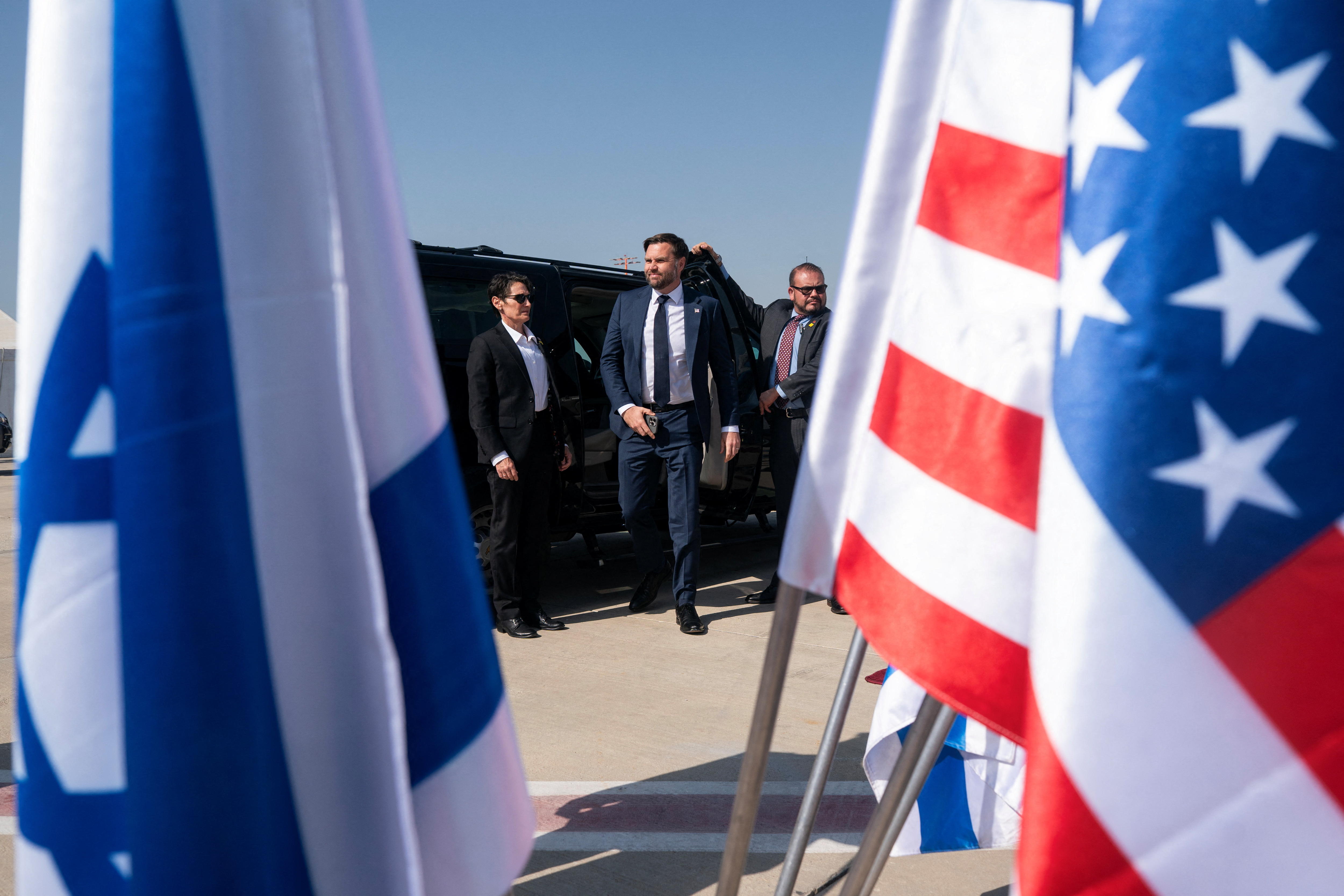 A man in suits steps out of a vehicle parked near Israeli and American flags.