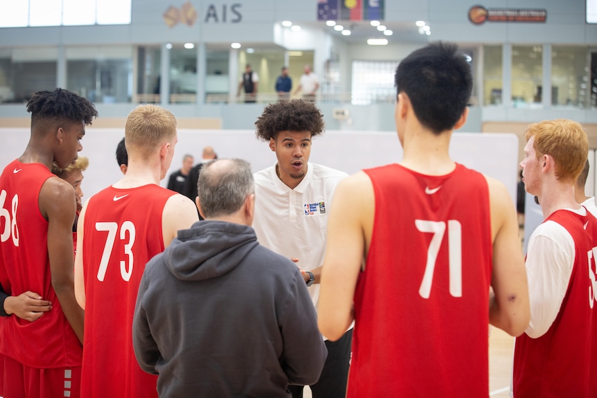 A man in a white shirt is surrounded by basketballers wearing red tops, who are all listening to him talk.