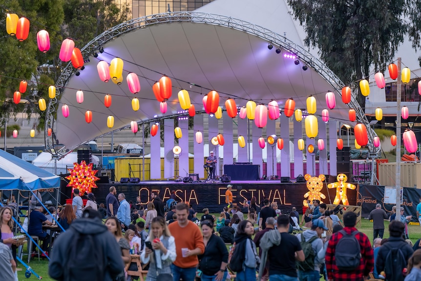 People stand under multicoloured Chinese lanterns strung up across a park, in front of a stage, which reads OzAsia Festival