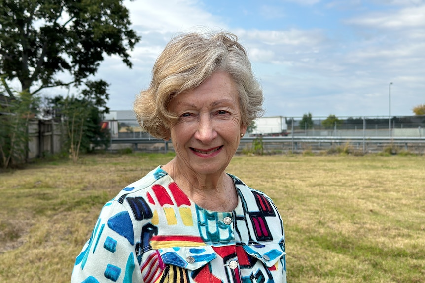 Eine ältere Frau lächelt in die Kamera und trägt ein farbenfrohes Hemd mit einem leeren Wohnblock im Hintergrund. 