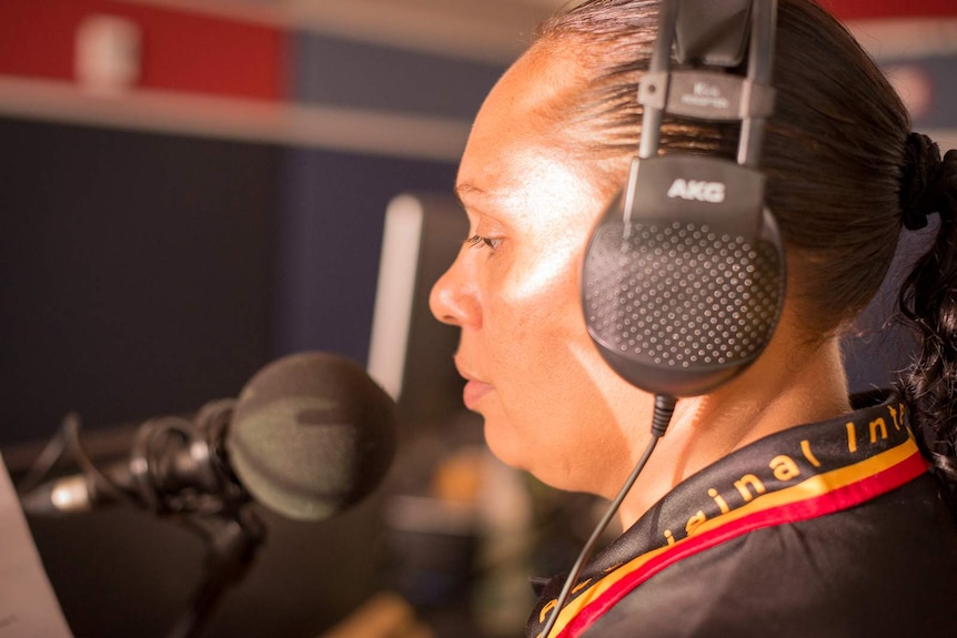 A woman wearing studio headphones speaks into a microphone as she reads from a script.