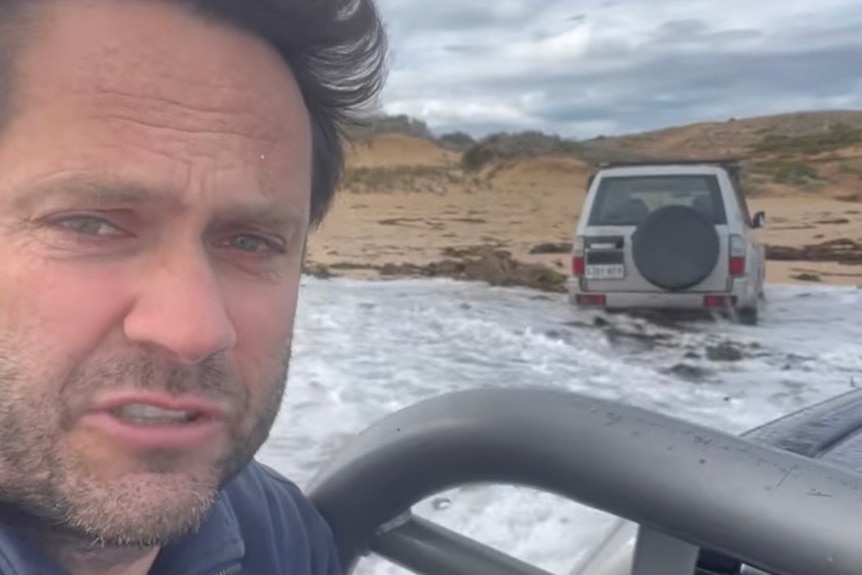 A man looks at the camera while sitting in the back of a ute that is bogged on the beach as waves wash in.