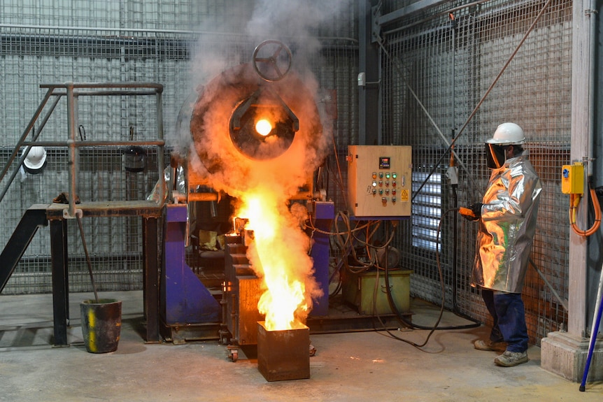 Un homme, vêtu d'un costume en argent métallisé, se tient à côté d'un grand chaudron versant de la boue enflammée dans des étagères empilées.