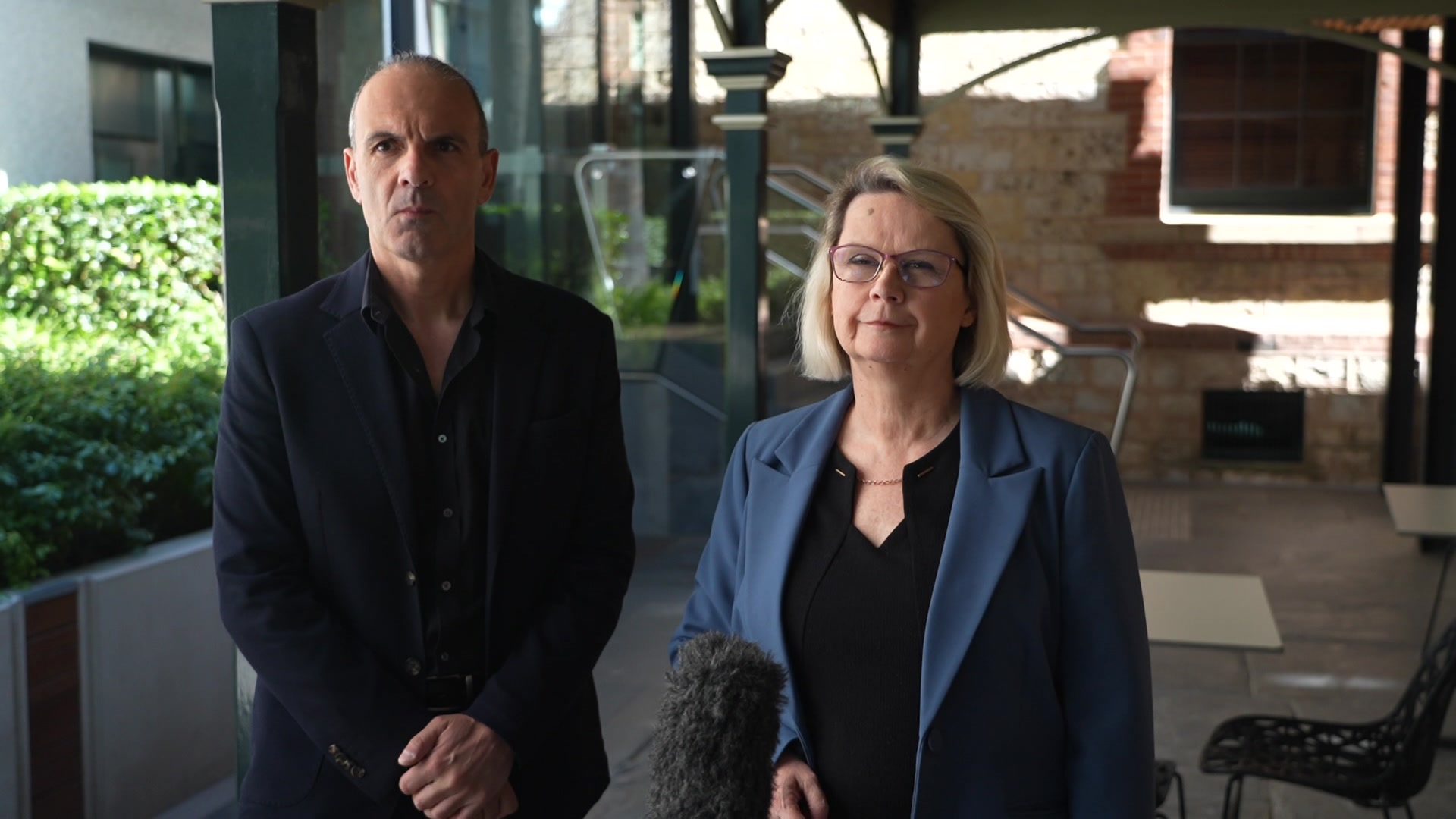 Um homem de terno escuro e uma mulher loira de paletó estão em uma área sombreada e conversam com a mídia.