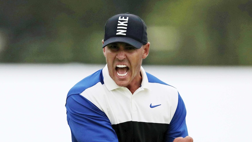 Brooks Koepka punches the air and screams in delight wearing a blue and white shirt and black hat