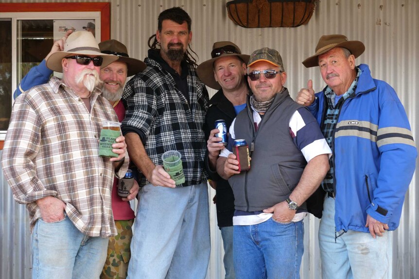 A group of men holding beers smile and give the thumbs up at the camera.