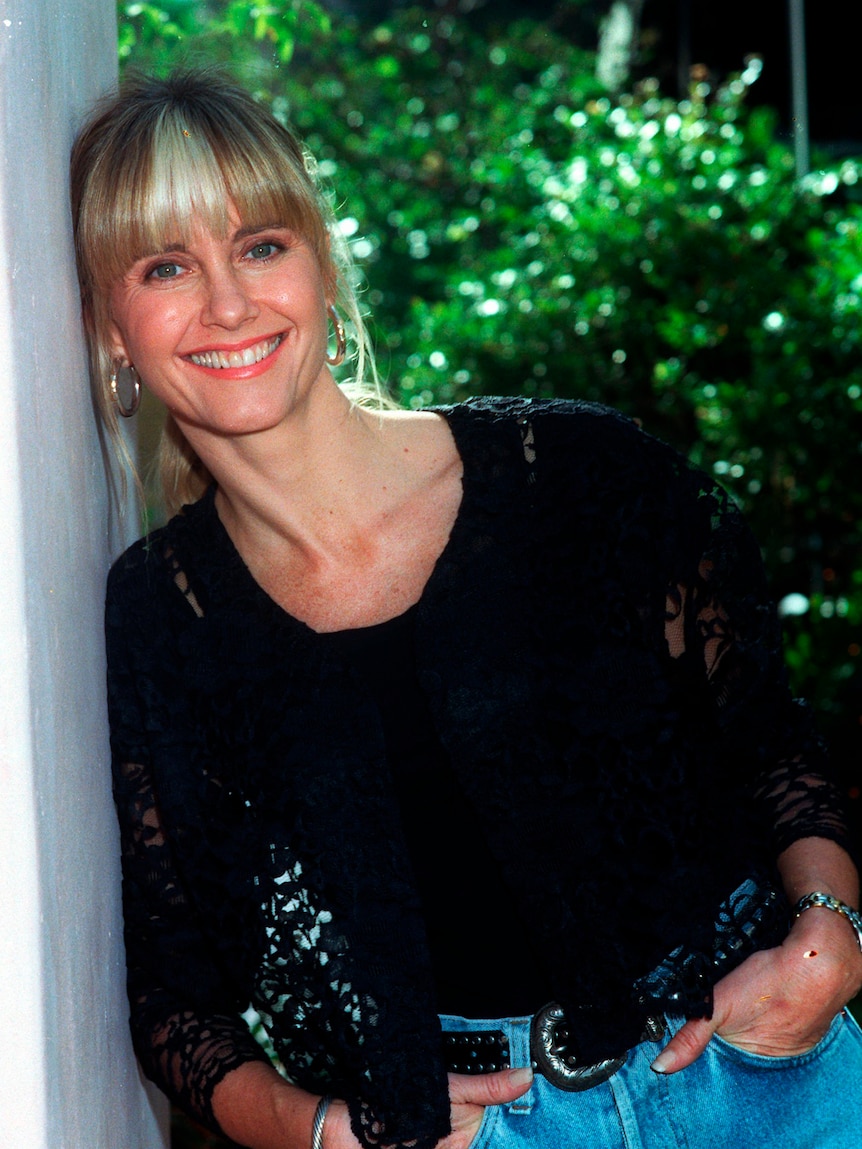 Olivia Newton-John leans against a wall with her hands in her pockets