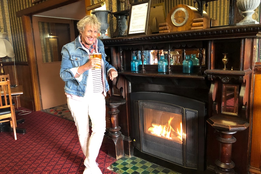 Woman standing next to a lit fireplace in an old fashioned pub, smiling and holding a beer. 
