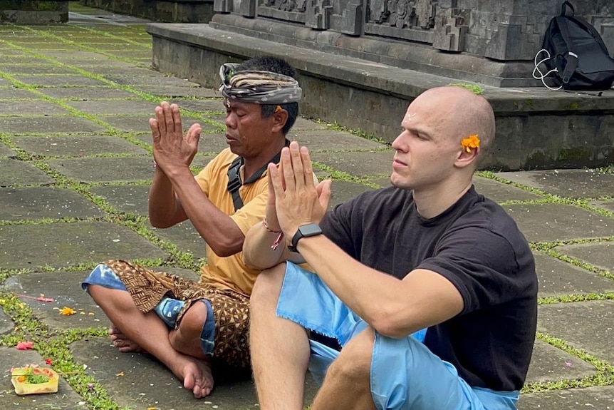 Two men sit on the paved ground, with their legs crossed and arms in prayer position. Both have their eyes closed