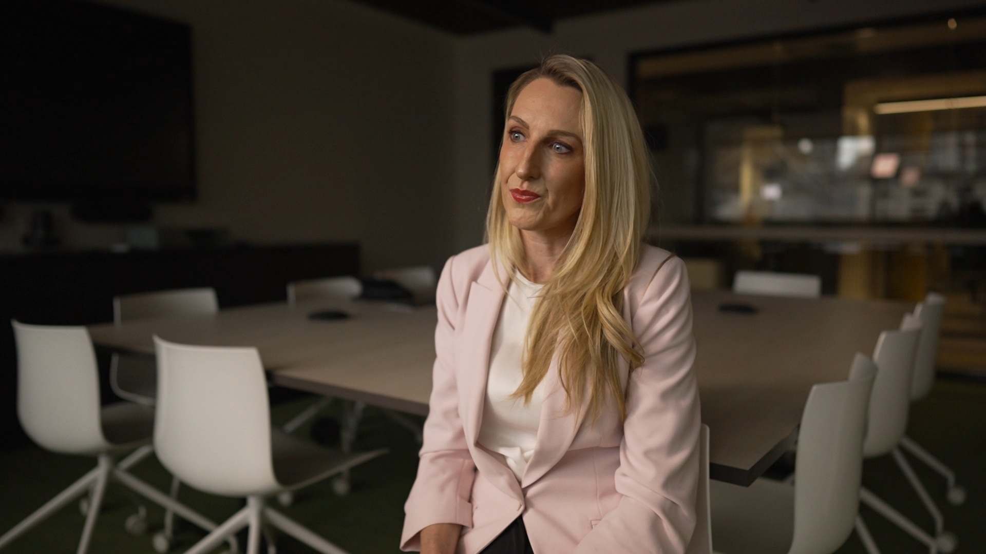 A middle aged woman with long blonde hair wearing a light pink blazer sits on a chair in a large meeting room.