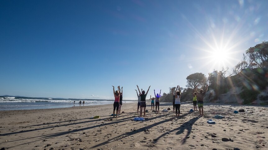 Yoga participants on a beach raise their arms above their heads, their shadows visible on the sand.