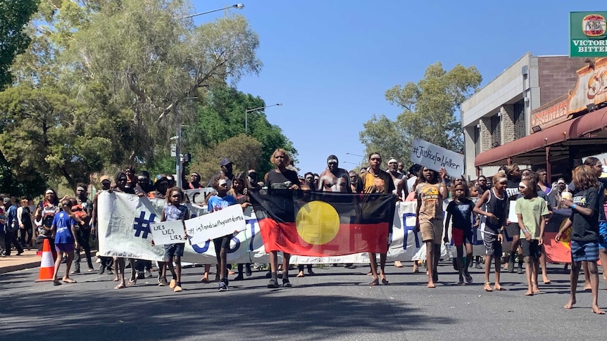 See from the front, a crowd of people walking down a street with young people at the front holding an Aboriginal flag