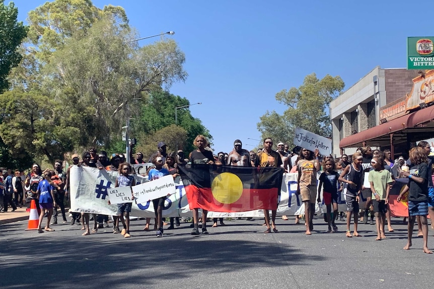 See from the front, a crowd of people walking down a street with young people at the front holding an Aboriginal flag