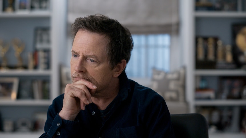 Actor Michael J. Fox, a man with short dark hair with grey patches, cups his chin in his hands, looking thoughtful.
