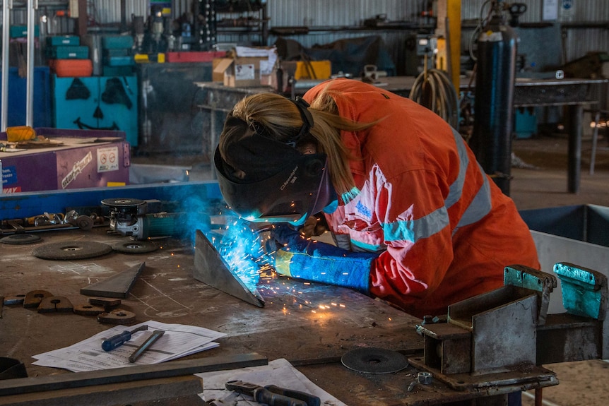 Woman with welding helmet on and fusing two pieces of metal together.